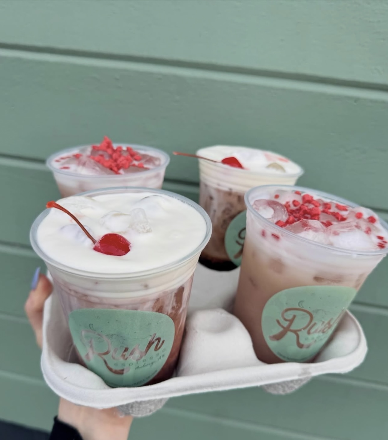 Tray of four iced Rush drinks against the sage siding